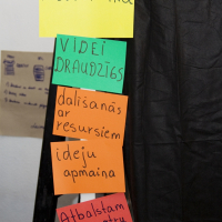 Five colorful signs are pinned vertically on a black fabric wall. The signs read (from top to bottom) "Modes talka" on yellow paper, "VIDEI DRAUDZĪGS" on green paper, "dalīsanās ar resursiem" on orange paper, "ideju apmaina" on red paper, and "Atbalstam viens otru prakt