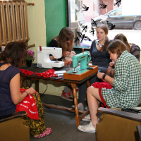 Five people are sitting and engaged in sewing activities around a wooden table. Two sewing machines, one white and one green, operate in different modes on the table. The room has green walls and a radiator. A window behind them shows a street with parked vehicles. Flowers and decorations are also visible in the room.