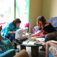 Four women are engaged in sewing activities in a cozy room. Two women are using a sewing machine on a round table, while another two are hand-sewing. They are surrounded by colorful fabrics and seated on comfortable chairs near a window, creating a relaxed, collaborative Modes Talka atmosphere.