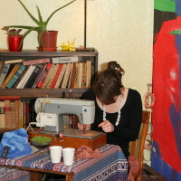 A woman sits at a wooden table using a sewing machine, surrounded by various fabrics and cups. The table is covered with a colorful, patterned cloth. Engaging in modes talka, she finds inspiration from the bookshelf filled with books and potted plants behind her. A vibrant, abstract painting decorates the wall as a lamp illuminates the space.