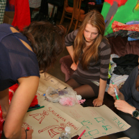 Three women are gathered around a table, engaging in collaborative drawing, or "talka." They are using markers on large sheets of paper filled with sketches of clothing. The scene is set in a colorful, busy environment with various fabrics and artworks illustrating different fashion modes in the background.
