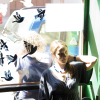 A woman sits by a sunlit window decorated with bird stickers. She has long hair tied in a low ponytail and is wearing a necklace and a dark top with short, loose sleeves. Next to her Modes Talka sewing machine, the bright sunlight casts bird silhouettes on her and the wall.