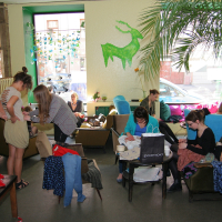 A lively room filled with people engaged in various activities. Some are standing, others are seated, all focused on tasks like sewing, drawing, or crafting using Modes talka techniques. The walls are adorned with green unicorn-themed decorations. A large potted plant and windows allow natural light to brighten the space.