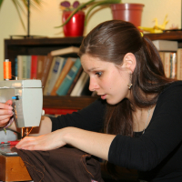 A young woman with long brown hair is using a light-colored sewing machine. She is focused, guiding brown fabric under the needle with one hand while adjusting settings using the various Modes with the other. The scene is indoors, with bookshelves and potted plants in the background, creating a cozy, lived-in feel.