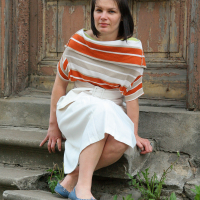 A woman with shoulder-length dark hair sits on an old, weathered stone step. She is wearing a striped top with orange, beige, and white colors, and a white skirt. Her blue flats add a touch of charm. The background features a worn-out wooden door with peeling paint, enhancing the rustic Modes talka atmosphere.