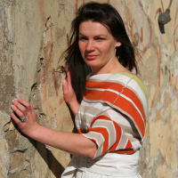 A woman with straight, dark hair poses against a weathered, textured wall with peeling paint. She wears an orange and white striped top and a white skirt, her left hand resting on the wall. Gazing directly at the camera in bright, natural sunlight, she embodies a modern mode talka vibe.