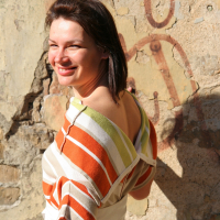 A smiling woman stands against a weathered wall with peeling paint and faded graffiti. She has shoulder-length dark hair and wears an off-the-shoulder striped top in orange, green, and white colors, paired with a white skirt. The sunlight casts subtle shadows, highlighting her cheerful expression as she enjoys her modes talka surroundings.