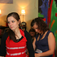 Two young women stand indoors, engrossed in different activities. The woman in the foreground wears a red and white tie-dye top over a black long-sleeve shirt, looking off to her right with a sense of modes talka. The woman behind her in a blue top focuses on something ahead. Warm lighting, visible lamps, and a colorful abstract painting add to the room's ambiance.