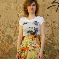 A person with shoulder-length hair stands against a worn, graffiti-marked wall. They wear a white T-shirt featuring a face with a butterfly and a patterned skirt with orange flowers and leaves. The wall, evocative of various artistic modes, has peeling paint revealing patches of brick and plaster.