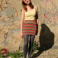 A woman with straight, light-brown hair stands against a weathered, graffiti-marked wall. She wears a yellow tank top, a brightly striped skirt, dark leggings, and grey flats with bows. Exuding casual Modes energy, she smiles slightly and poses with her head tilted to one side. Small plants grow near her feet.