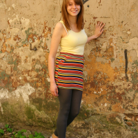 A young woman with light brown hair, wearing a yellow sleeveless top, a striped mini skirt, black leggings, and grey shoes, is standing against a weathered, graffiti-covered wall. She has a relaxed smile and her right hand is touching the wall. The ground is covered in small stones and greenery; she looks like she's ready for some Modes talka.