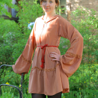 A woman with short brown hair stands outside in front of greenery and a building. She is wearing a loose-fitting peach-colored dress with long, billowy sleeves, cinched at the waist with a red cord. With one hand on her hip and the other resting on the back of a metal chair, she exudes modes talka.