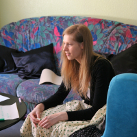 A woman with long blonde hair is sitting on a blue chair, wearing a black top and a white polka-dotted skirt. She is smiling and looking to her right. In the living room, with its colorful floral-patterned couch and black cushions behind her, sits a table scattered with papers from her latest Modes talka.