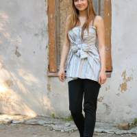 A young woman with long hair stands in front of a weathered wall with peeling paint and a boarded-up window. She is wearing a striped sleeveless top tied in front, black pants, and black shoes. A small flower accessory is clipped to her hair. With a slight smile, she could be the muse for Modes Talka.