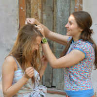 Two young women with long hair stand in front of a weathered wall. The woman on the right, wearing a floral shirt and a green wristwatch, is braiding the hair of the woman on the left, who wears a light striped top and looks down, smiling. They both appear to be enjoying the moment in quiet modes talka.