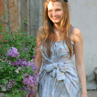 A young woman with long, light brown hair stands outdoors next to a blooming lilac bush. She wears a sleeveless, striped top with a large bow at the waist and black pants. Smiling as she gently holds a branch of the lilac bush, she exudes an effortless charm amidst the wooden and concrete elements in the background.