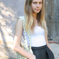 A young woman with long blonde hair stands outdoors, wearing a white strapless top and a black skirt with white polka dots. She has a matching polka dot hair accessory and a green polka dot bag. The background features a rustic, weathered wooden door and sunlit wall. Modes talka, she has her hand in her skirt pocket.