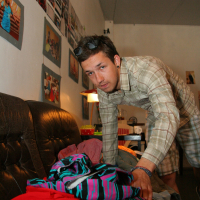 A person wearing a plaid shirt and plaid shorts leans over a sofa, sorting through a pile of colorful clothes. The room is decorated with various photographs and posters on the walls. There is a lamp on a small table behind the person, its light showcasing their modes talka collection.