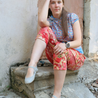 A young woman with long, brown hair, wearing a blue floral shirt and red floral pants, sits in a talka mood on a small concrete step against a weathered wall and door. She rests her left elbow on her knee and her head on her hand, looking into the camera. She wears gray flats and a wristwatch.