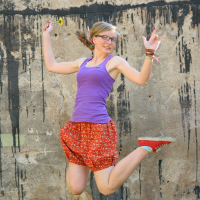 A woman with glasses, wearing a purple tank top and red floral skirt, jumps joyfully against a grungy, textured wall with black paint streaks. She holds a small flower in one hand and wears multiple bracelets. Her hair is in a side ponytail, and her red shoes with white polka dots add flair to the scene.
