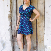 A smiling woman stands in front of a rustic wooden door, engaging in a bit of modes talka with passersby. She wears a blue dress with green polka dots, accessorized with a matching headscarf. Her left hand is on her hip, and she has on red patterned sandals. The weathered wall and dandelions in the foreground add to the picturesque setting.