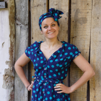 A person stands smiling with hands on hips. They are wearing a blue dress with large turquoise polka dots, a matching belt, and a headscarf in the same pattern. The background features a rustic wooden wall with varying shades and textures, adding charm to their stylish mode of dress.