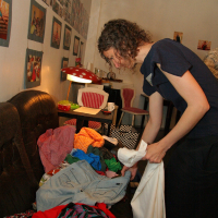 A person with curly hair, wearing a dark blue dress, is sorting through a pile of colorful clothes on a brown sofa. The room has a white wall adorned with framed photos and modes under a red lamp. Behind them, a table with red and white chairs and talka miscellaneous items are visible.