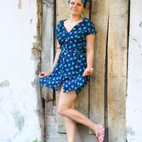 A woman stands against a rustic wooden door, wearing a blue dress with green polka dots and a matching headscarf by Modes Talka. She has her left foot raised, displaying floral-patterned shoes. Her hands are playfully tugging at her dress as she smiles warmly. The background features weathered wood and stone.
