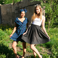 Two young women stand in a sunlit, overgrown garden. The woman on the left wears a blue polka-dot dress and headscarf, smiling. The woman on the right wears a white tank top and black polka-dot skirt, also smiling. Both hold onto their skirts, creating an airy, playful atmosphere amidst the green talka.