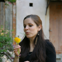 A woman with long brown hair wearing a black top is standing outdoors and blowing on a dandelion. The background shows an old building with a wooden door and greenery, including a flowering tree to the left. Her expression appears focused on the modes talka as the dandelion seeds disperse into the air.