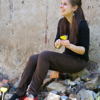 A woman sitting on a pile of bricks and rubble, smiling and holding a yellow dandelion. She has long dark hair and is wearing a black long-sleeve shirt, dark pants, and red shoes. The weathered concrete wall forms the background as she’s surrounded by green grass with more dandelions—modes talka unfolding around her.