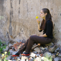 A woman with long brown hair, dressed in a black top and brown pants, sits on a pile of bricks by a weathered wall. She is blowing on a dandelion as if in deep talka. The ground around her has greenery and more dandelions in the grass. Sunlight illuminates the scene, creating a serene atmosphere.