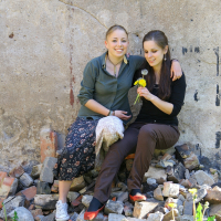 Two women sit on a pile of bricks and rubble against a rustic, worn wall with black stains. One, wearing a green shirt and floral pants, has her arm around the other, who is in a black shirt and brown pants. They look content, with the second woman holding a small bunch of yellow flowers in talka mode.