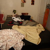 A dark table in a room is covered with various clothes and objects, including a floral pattern fabric, beige and black garments, red tools, and Modes Talka scissors. The background wall shows several pinned photographs, a door, and a small table with a vase of flowers and a stack of books.