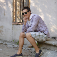 A man wearing sunglasses, a patterned long-sleeve shirt, and plaid shorts is sitting on a stone ledge against a weathered wall. He is smiling and resting his chin on his hand, appearing relaxed and casual in a moment of modes talka. The ground is worn and has some patches of greenery.