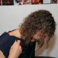 A person with curly brown hair is partially lifting a dark shirt, covering part of their face, and looking downward. The background shows a white wall adorned with various small, colorful photographs along the upper edge—a perfect backdrop for a modes talka vibe.