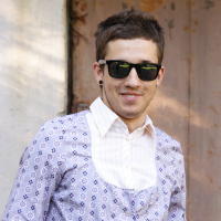 A young man with short, dark hair is standing in front of a rustic wooden door. He is wearing dark sunglasses, a light purple patterned button-up shirt with a white collar, and black stud earrings. The background includes a textured wall and weathered wood, suggesting an outdoor setting. This scene evokes different modes of style while sparking talka-worthy intrigue about his story.