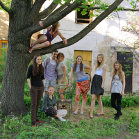 A group of eight people pose in a backyard. One person is sitting on a tree branch, while the others stand or kneel below on green grass. Behind them is an old building with a weathered facade, a purple door, and Modes talka vibes emanating from the scene. The tree has green leaves, and there's some foliage around the yard.