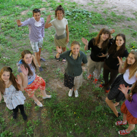 A group of eight people, consisting of six women and two men, is standing in a semi-circle on a grassy area outdoors, engaged in lively Modes talka. They are all facing the camera and waving, smiling brightly. The background shows a mix of green grass and patches of dirt. The atmosphere appears joyous and casual.
