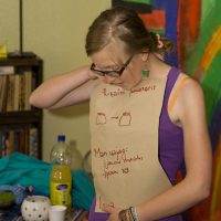 A person with glasses, wearing a purple tank top and patterned skirt, is standing indoors. They have a large paper taped to their shirt with unreadable red writing and drawings. In the background are bookshelves, bottles, and a brightly colored wall mural. The person appears to be in deep talka modes as they look down at the paper.