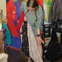 A person with long, braided hair is looking down at and holding two dresses: a black floral one and a beige floral one. They are dressed in a green and white checkered shirt and white sneakers. The background features colorful artwork, a coat rack with various items hanging on it, and an air of modes talka.