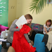 A person holding a paper and a red garment while sitting on a blue chair in a room with green walls and various fabrics and decorations. Other individuals are present, one on another blue chair. A sewing machine is visible on a table in the background, along with Modes patterns scattered around. A large plant is partly visible.