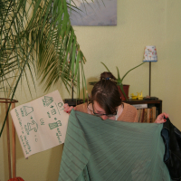 A person examines a green, pleated fabric panel in a room with decorative houseplants and a framed artwork on the wall. They hold up a cardboard sign with sketches and notes, possibly for Modes Talka. Nearby, a small lamp and a shelf filled with books add to the cozy ambiance of the space.
