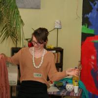 A person wearing a light brown sweater with a "Linda" name tag, white beaded necklace, and eyeglasses holds a peach-colored cloth in one hand and a clear bottle with yellow liquid in the other. They stand in Modes Talka, surrounded by vibrant abstract painting, bookshelves, and various decorations.