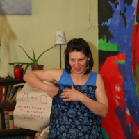 A woman with short dark hair, wearing a blue patterned dress and turquoise earrings, is standing indoors near a bookshelf and a large colorful wall painting. Sporting a cloth bag with a sketch and "Modes talka" text on it, she partially covers her outfit as various houseplants adorn the space.