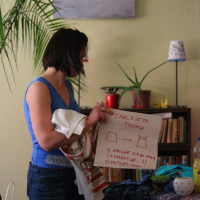 A woman in a blue tank top holds a whiteboard with text and drawings. Behind her are shelves filled with books and various items. A framed painting, a potted plant, and a lamp with a patterned shade decorate the space. Engaging in presentation modes, she appears to be teaching or giving a talka.