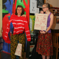 Two women standing indoors, dressed in colorful, patterned skirts and tops. One holds a red and white striped shirt, smiling. The other showcases a drawing with text and illustrations. The background features a vibrant, abstract wall mural and various posters beside a wooden chair and table—creating lively talka modes.
