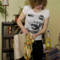 A woman with shoulder-length hair in a white T-shirt displaying a face print and black pants stands in a room holding a fabric with colorful patterns. Behind her is a bookshelf filled with various items, and on the table draped in talka-patterned cloth is an empty bottle. The wall is light green.
