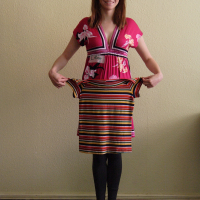 A woman with shoulder-length hair, wearing a pink floral dress over black leggings and grey flats, is standing indoors against a beige wall. She is smiling and facing the camera while holding up a colorful striped dress, showcasing her vibrant choice in modes talka.