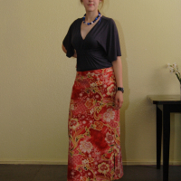 A woman stands indoors against a light yellow wall, wearing a dark gray top and a long, floral-patterned red skirt. She also has a blue beaded necklace, a wristwatch, and white shoes. Behind her, there is a framed painting on the wall and a small table with white flowers in full bloom—a tranquil mode of talka.
